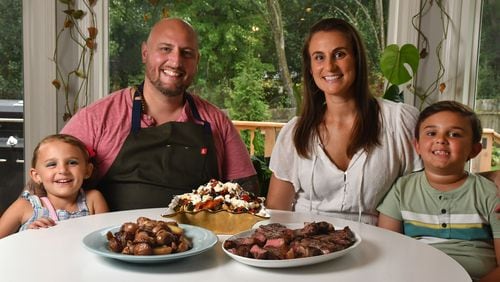 Chef Pat Pascarella sits with his wife, Megan, daughter Sophia, 5, and son Colin, 7, in the family's newly purchased Roswell home. The family is shown with one of Pascarella's favorite at-home dishes, New York Strip Steaks with Ciambotta Salad. There's also a bowl of grilled potatoes with onions because the kids want carbs, he says. (Styling by Pat Pascarella / Chris Hunt for the AJC)